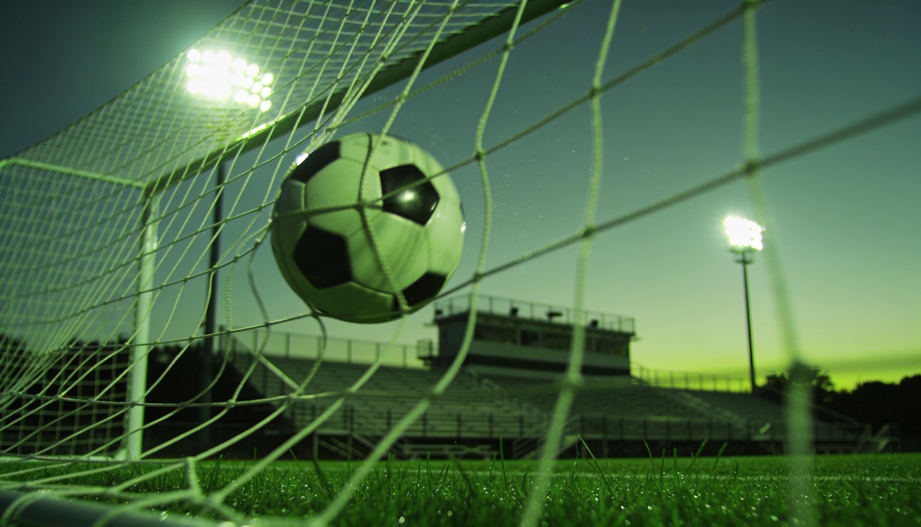 Soccer ball hitting the back of the net at an American stadium under the lights at dusk