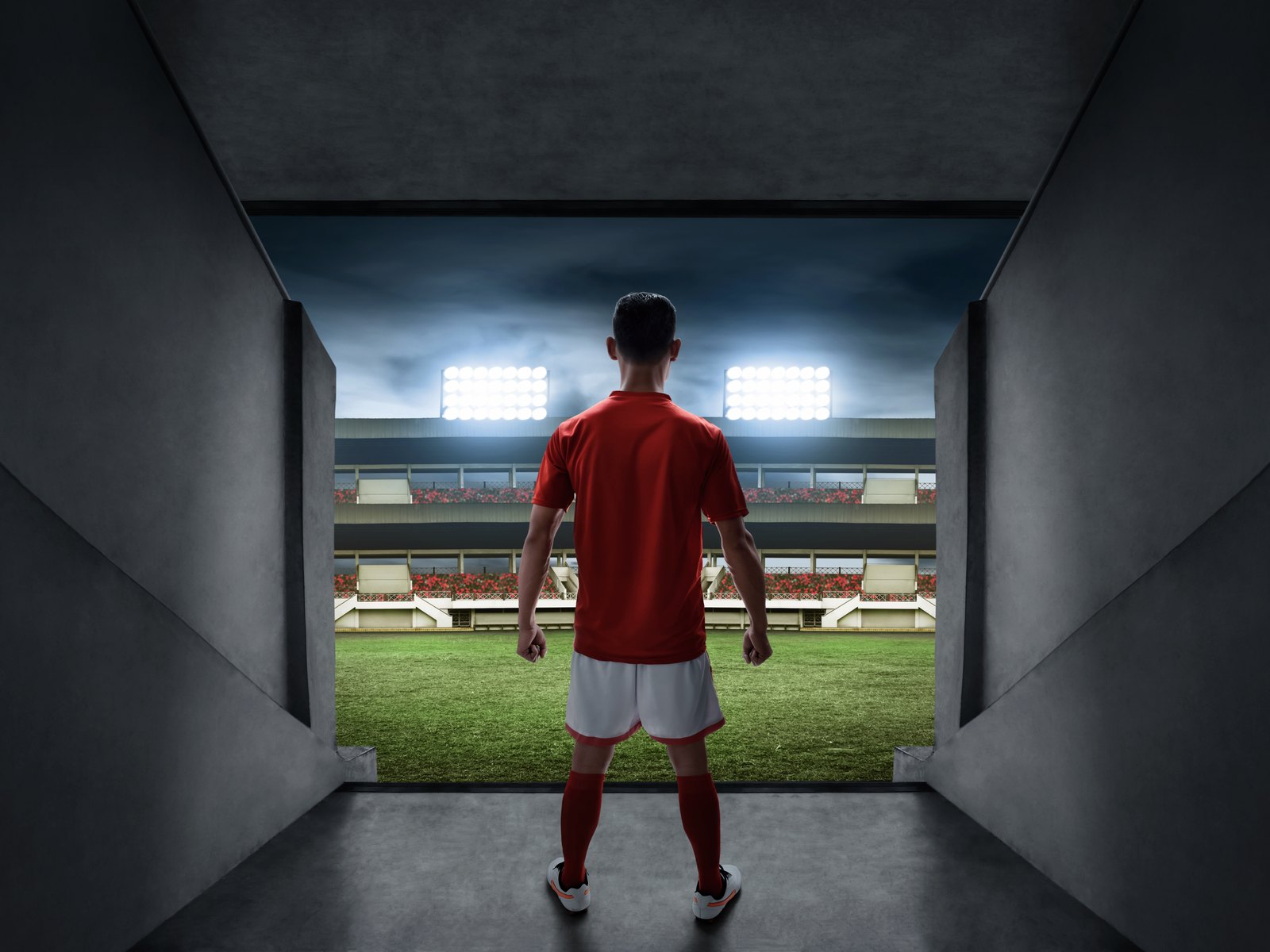 Young soccer player standing at the mouth of the tunnel, looking out onto a floodlit stadium pitch
